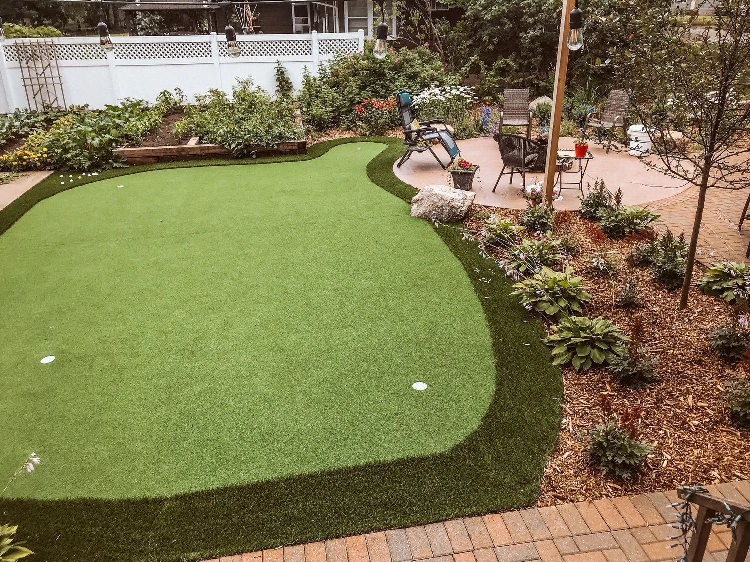 Backyard putting green surrounded by plants, bordering a patio with seating.