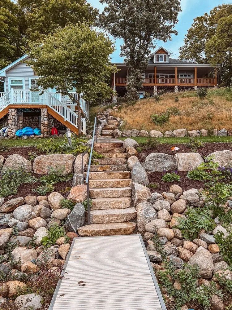 Stone steps lead uphill to a house with a porch, overlooking a waterfront and a smaller gray cottage.