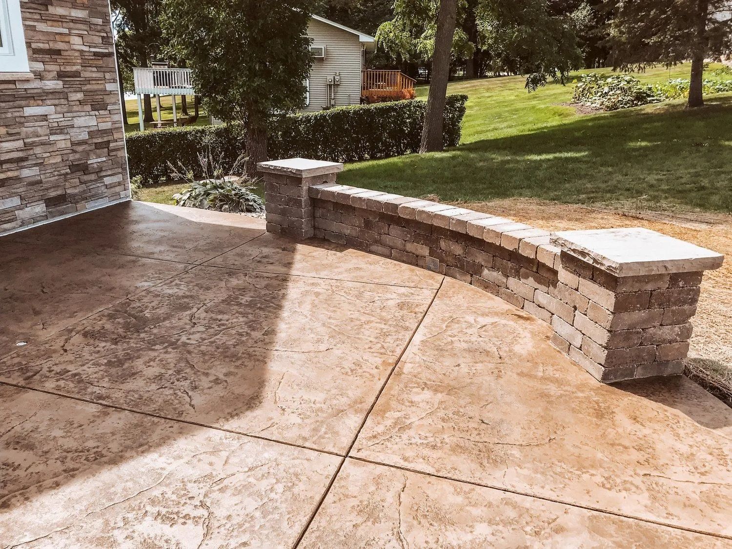 Brick curved retaining wall on a stamped concrete patio, next to a building with stone siding, sunny day.