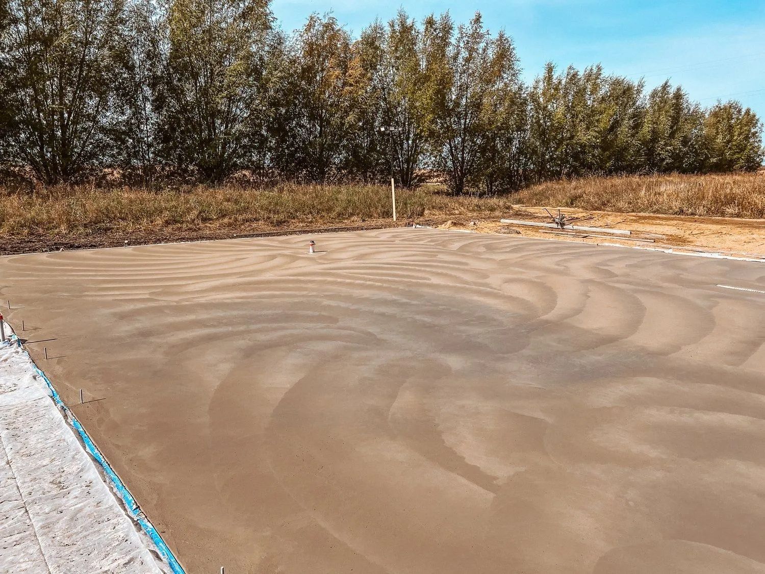 Freshly poured concrete slab in an open field, with swirl marks. Trees and blue sky in background.