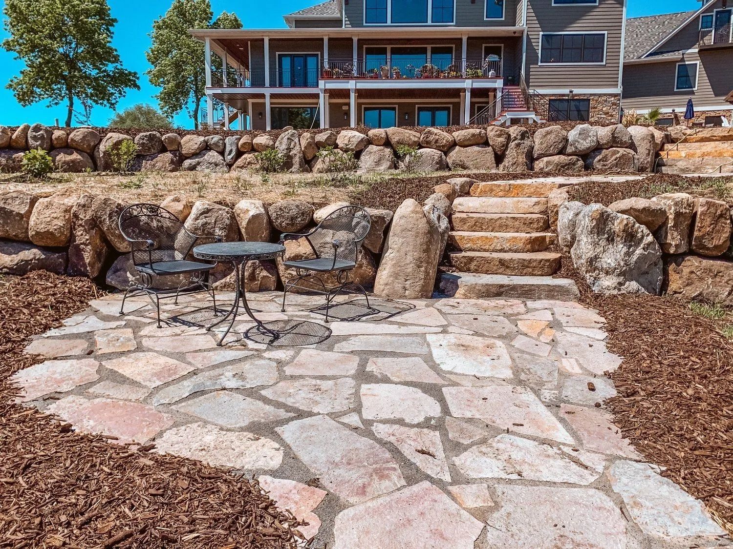 Stone patio with bistro set, steps, and rock retaining walls in front of a house.