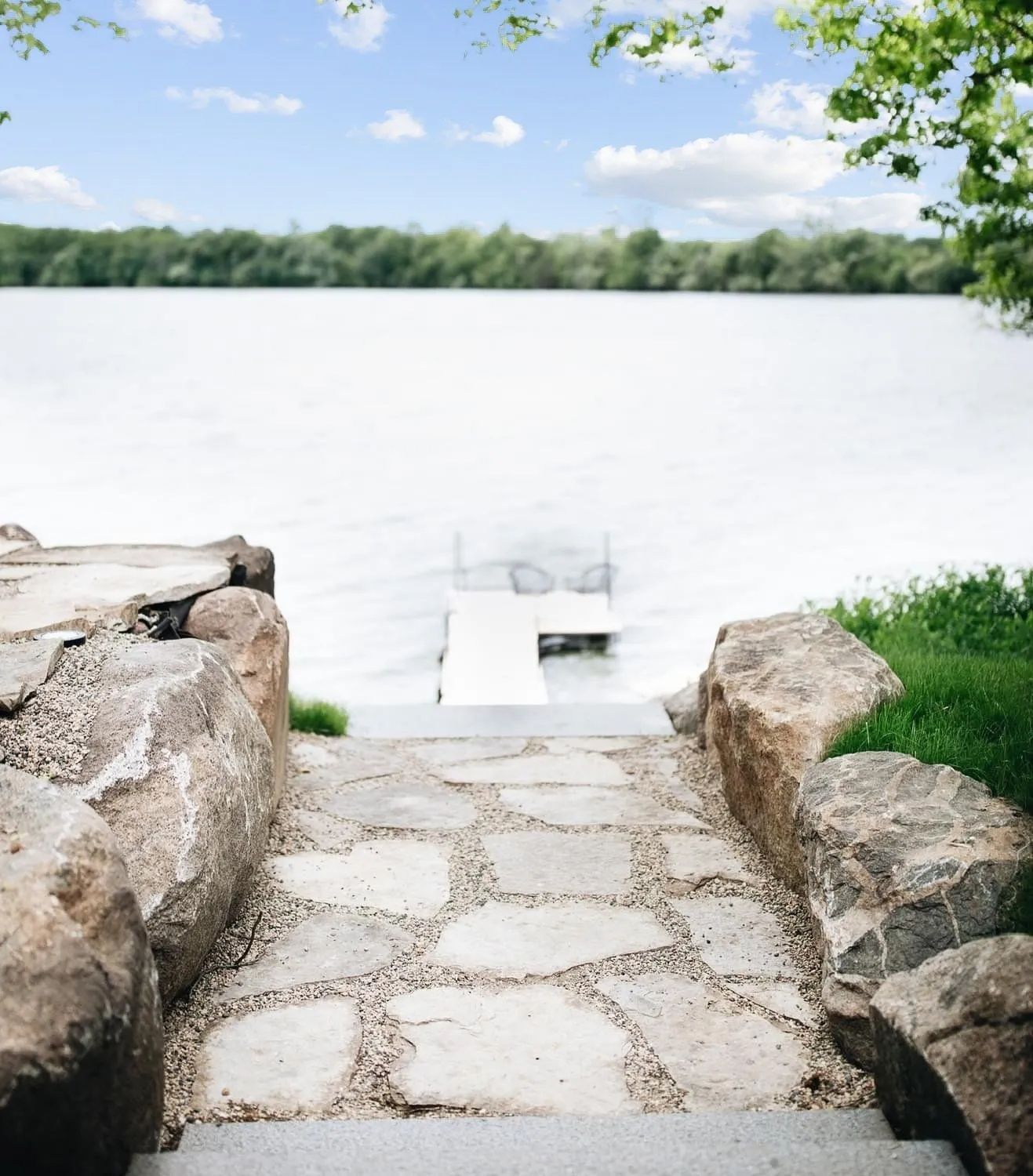 Stone steps leading to a white dock on a calm lake, framed by large rocks and green grass.