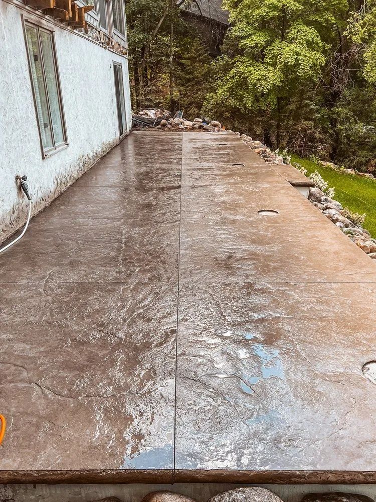 Concrete patio, half cleaned with pressure washer, near a house with lush greenery.
