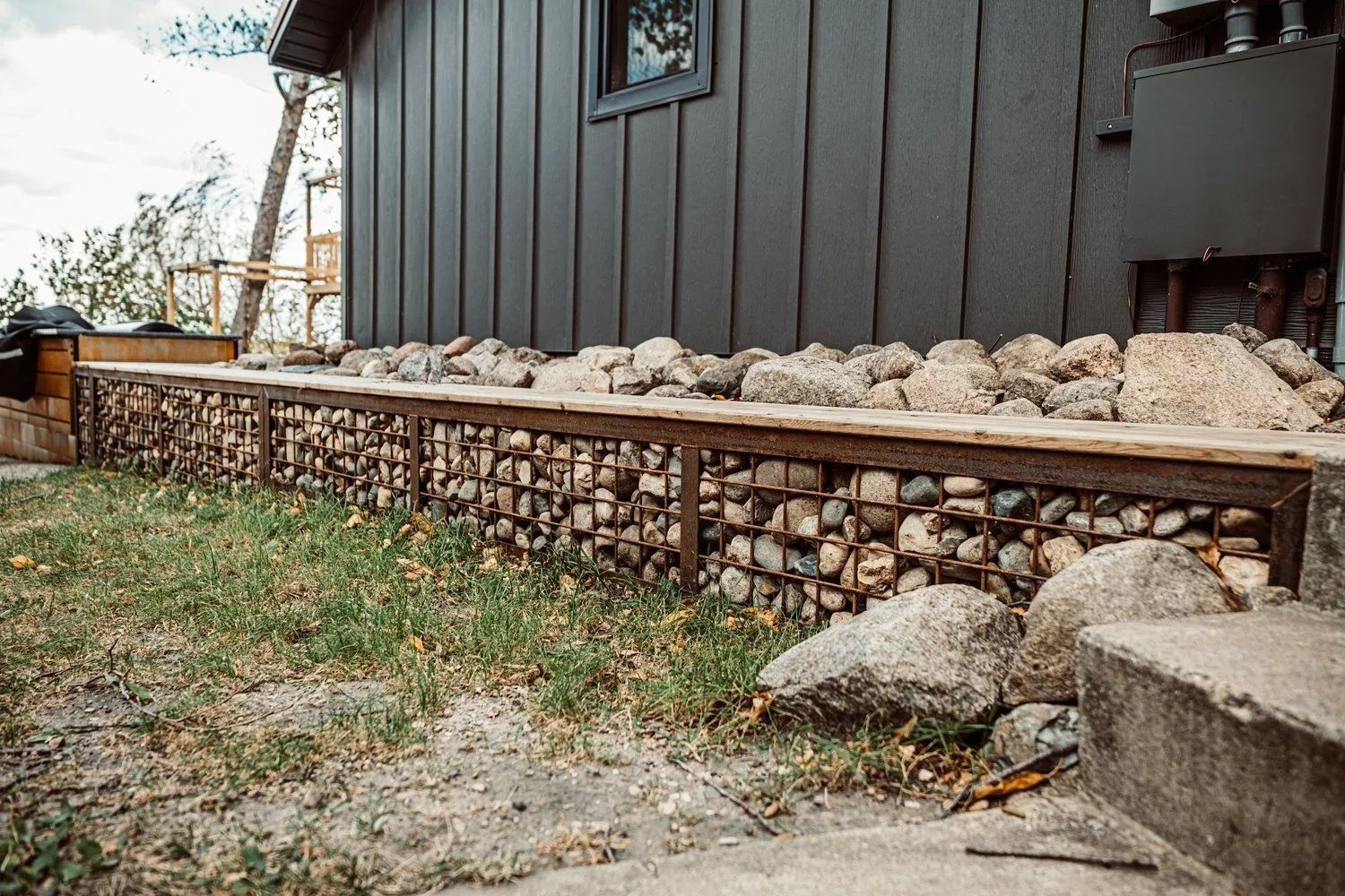 Exterior view of a dark gray building with a rock-filled gabion wall and wooden top, next to green grass.