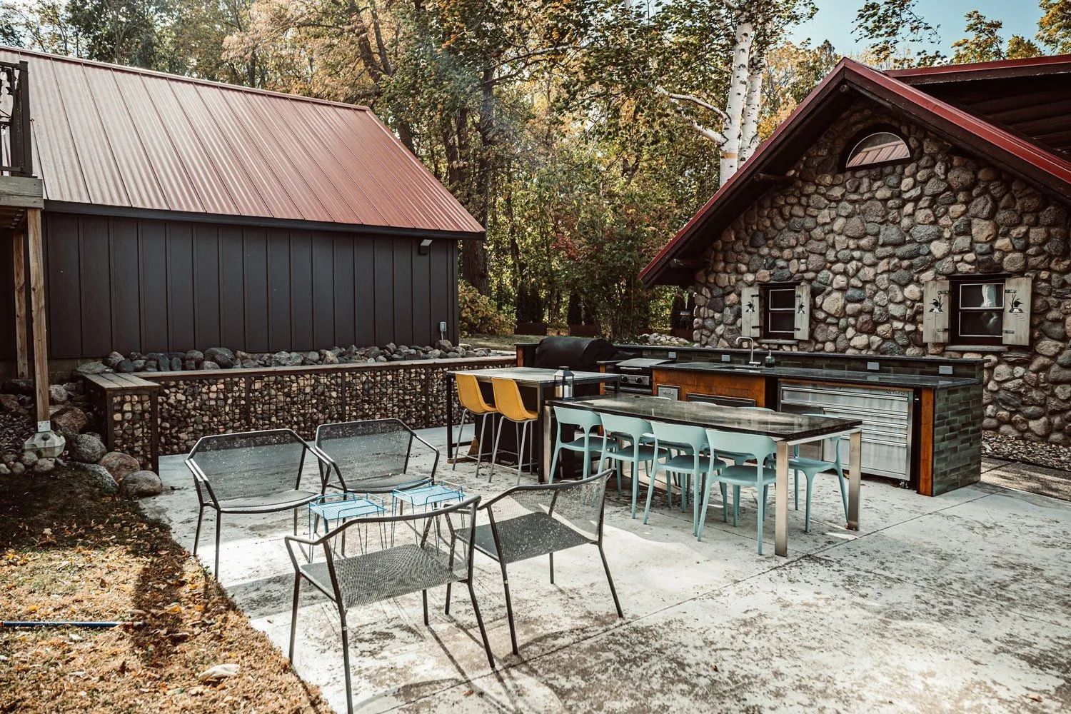 Outdoor patio with stone building, dining tables, and metal chairs, set in a wooded area.