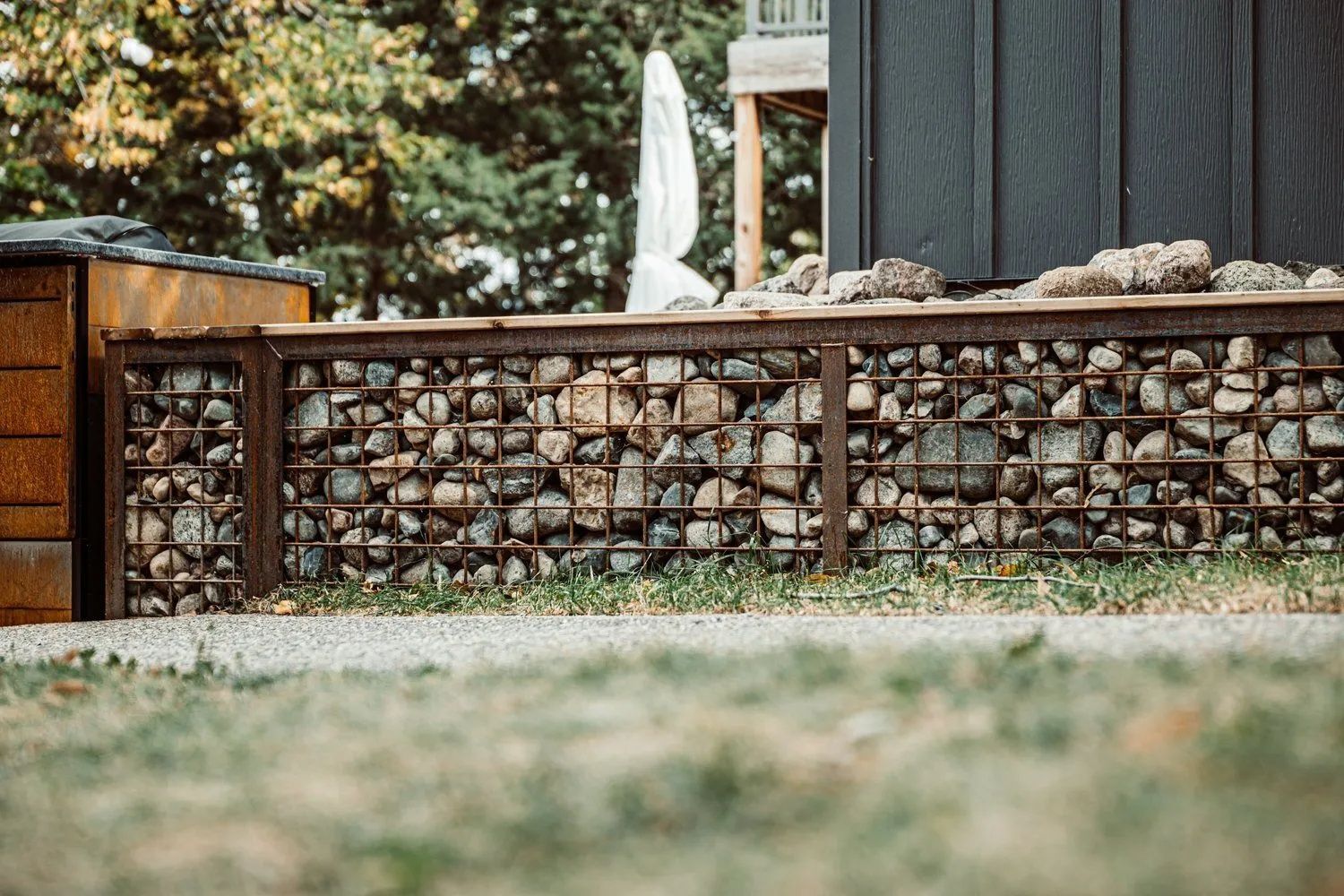 A rock-filled gabion wall with metal framing. Grass in the foreground and a wooden building in the background.