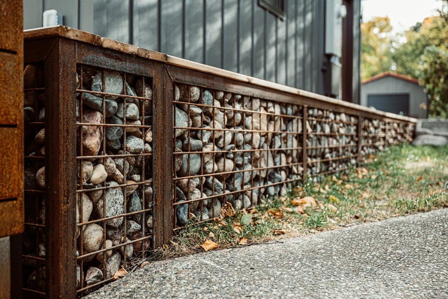 Stone and wire mesh retaining wall with wooden top along a concrete path.