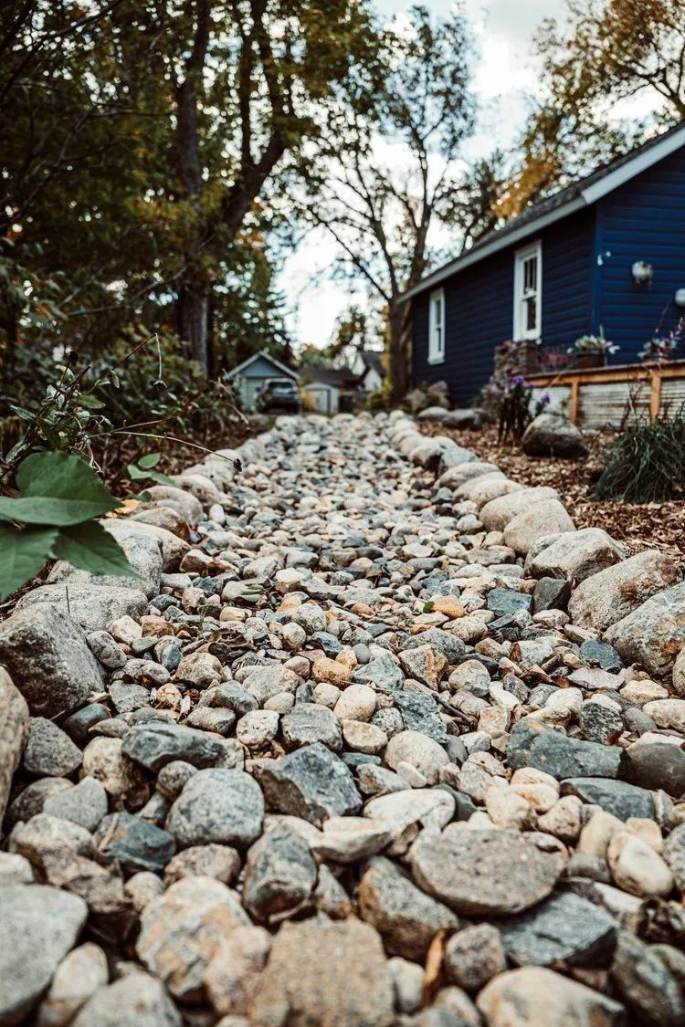 Gravel-filled dry creek bed leads toward a blue house with trees in the background.