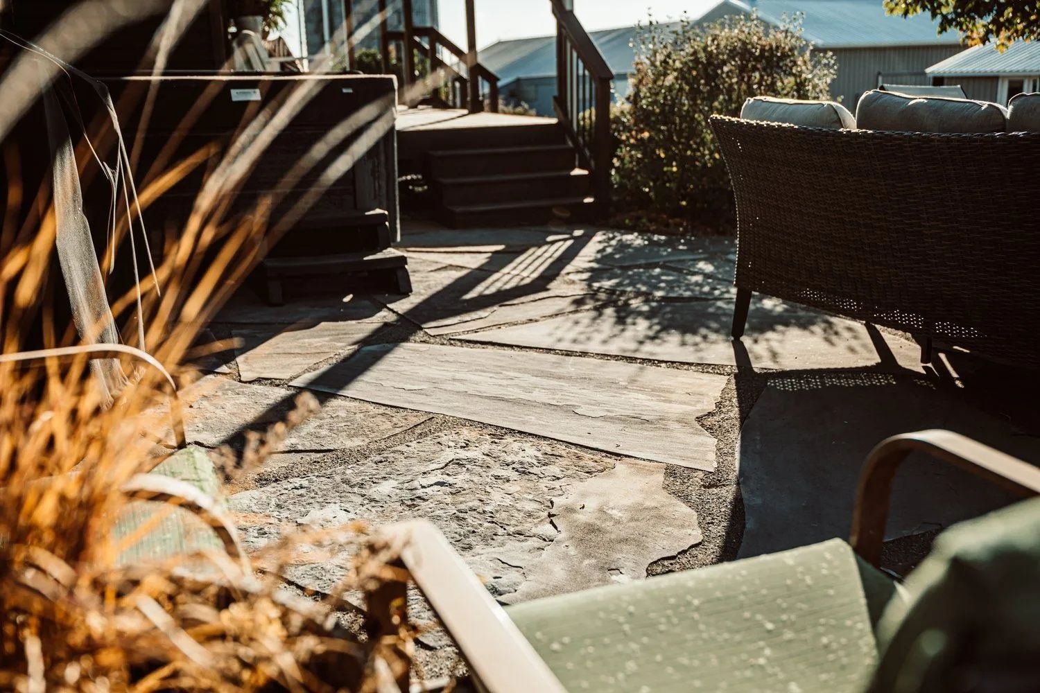 Patio with stone pavers, wicker furniture, and steps leading to a deck; sunlight and shadows.