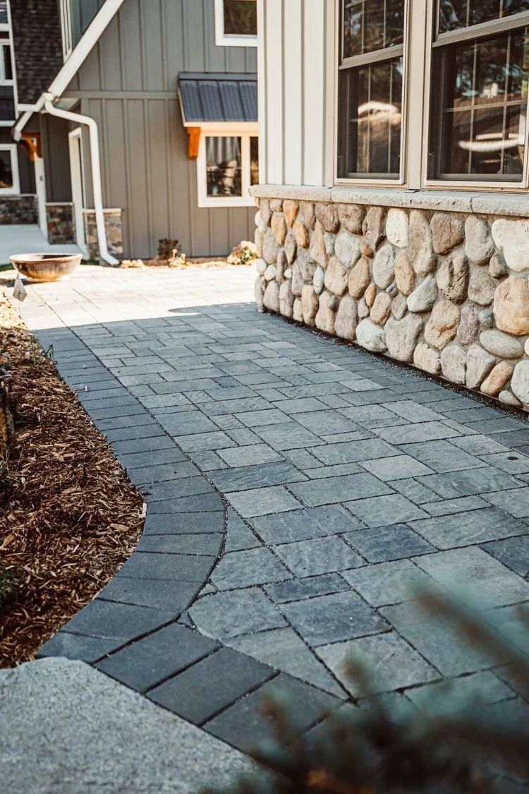 Stone pathway leading to a house with stone and wood siding, bordered by mulch and greenery.
