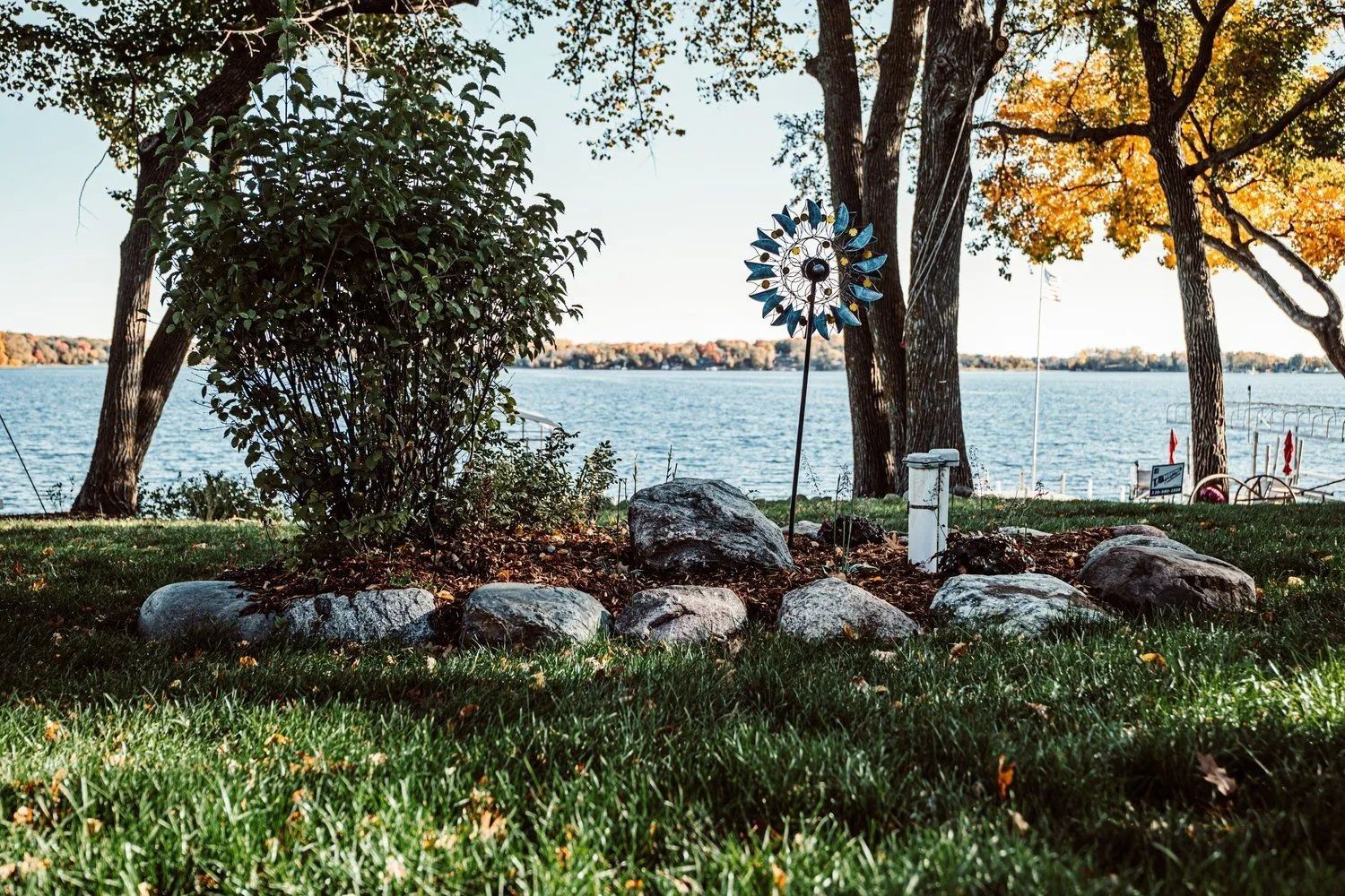Lakeside garden bed with large rocks, greenery, and a blue pinwheel; trees frame the water in the background.