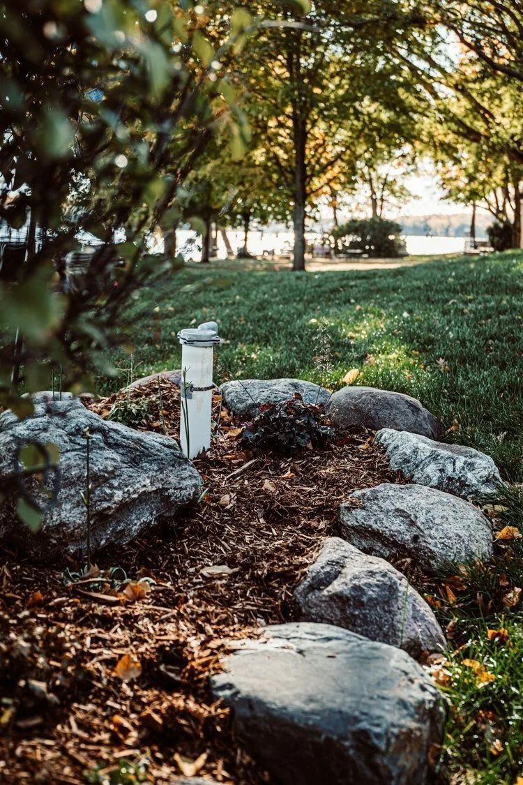 Landscaped garden bed with rocks, mulch, and a white pipe against a grassy hill with trees.