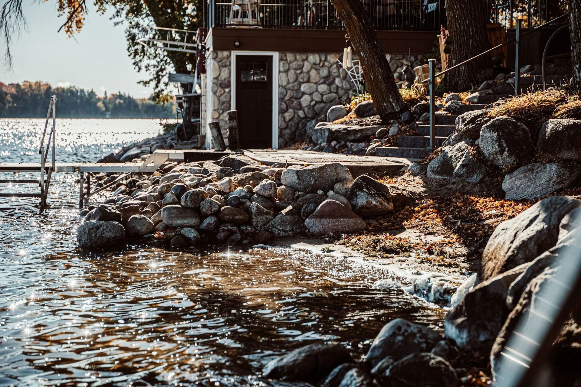 Lakeside stone building with a dark door, rock shoreline, and dock on a sunny day.