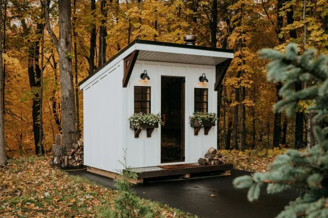 White cabin with a dark door, small windows, and flower boxes; set in autumn woods.