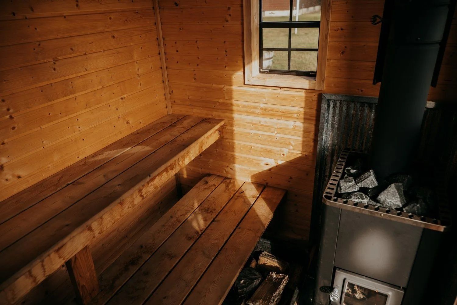 Wooden sauna interior with benches, stove with hot stones, and a small window.