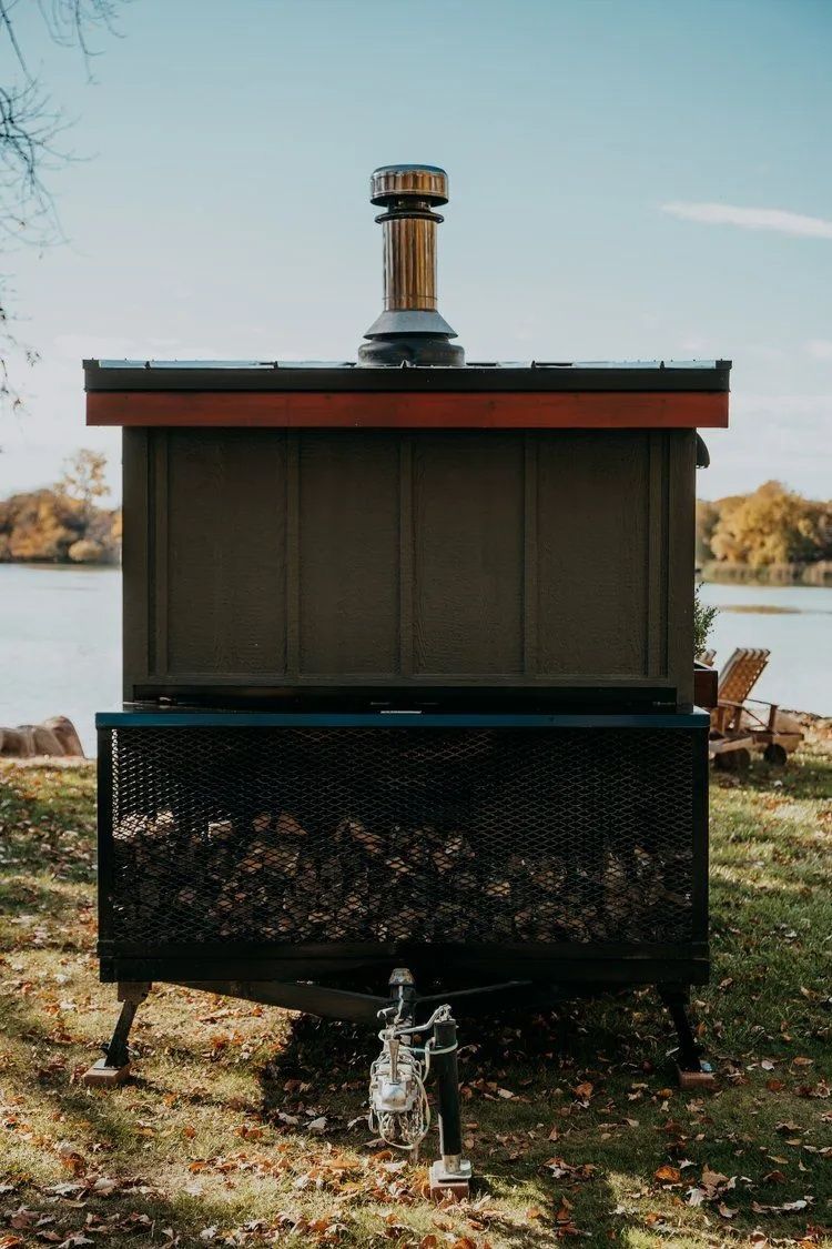 Outdoor wood-fired pizza oven with chimney. Metal structure, dark green sides, on grassy ground by water.