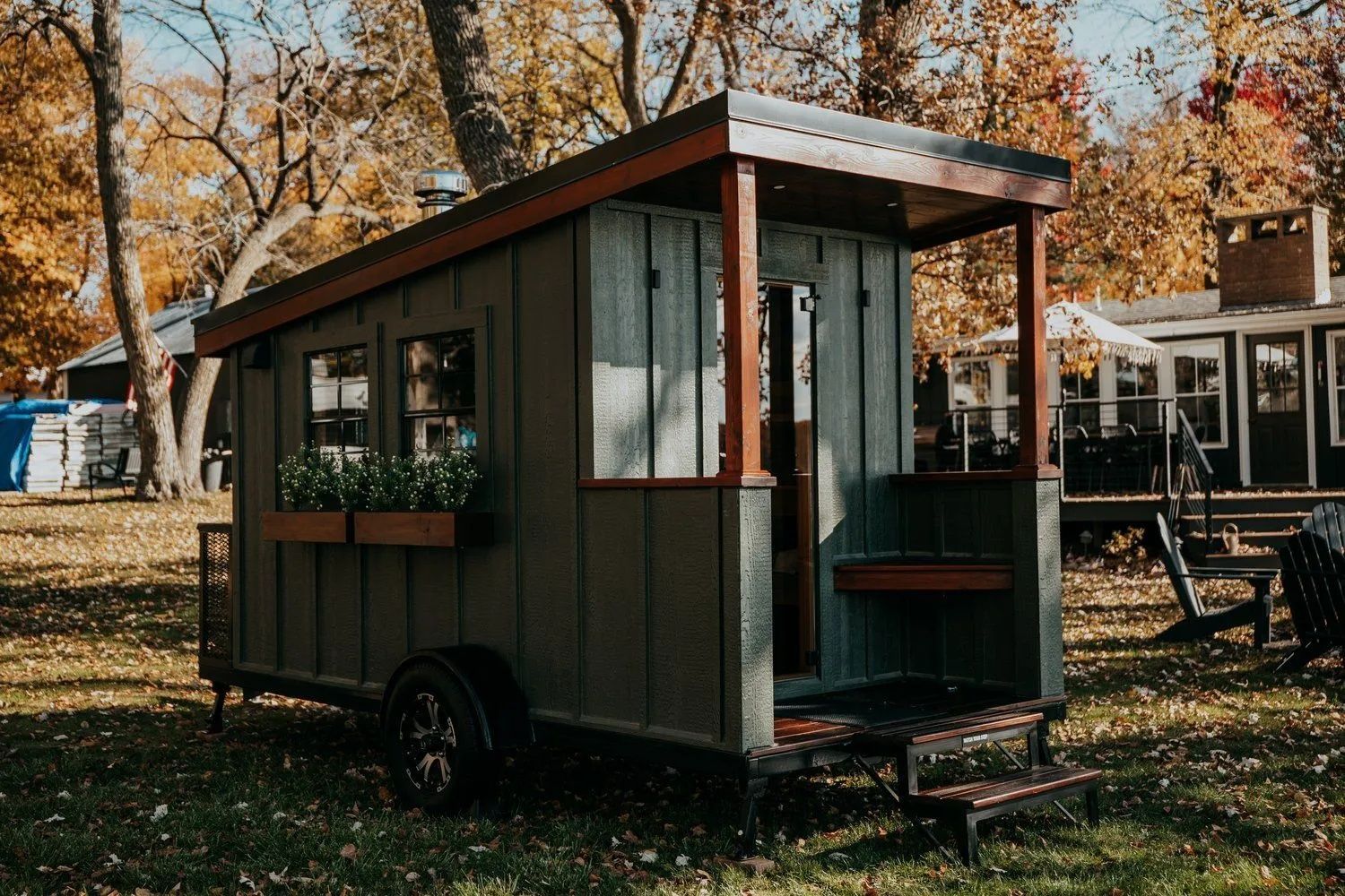 A small, green cabin on wheels with a porch and steps, set in a grassy yard in autumn.