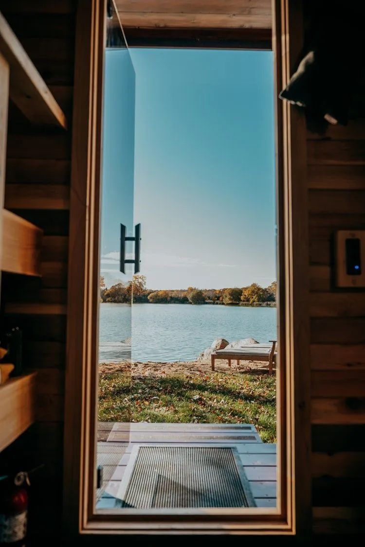 View of a lake through a tall window in a wooden cabin. Sunny day, blue sky, grassy bank.