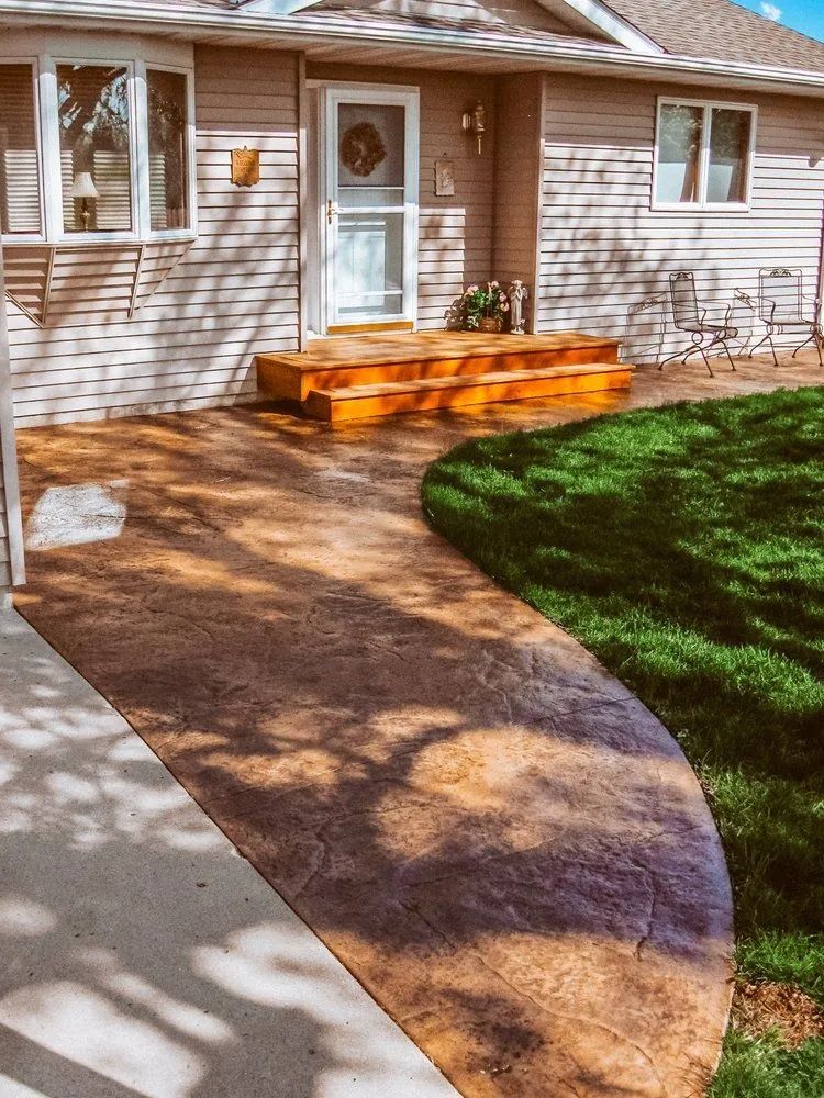 Exterior view of a house with a curved stained concrete pathway leading to a wooden front porch with steps, and green lawn.