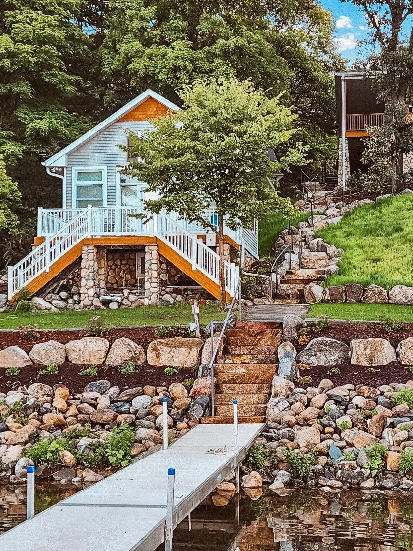 Cottage on a lake with a dock, steps, and rock retaining walls. Blue chairs on deck.