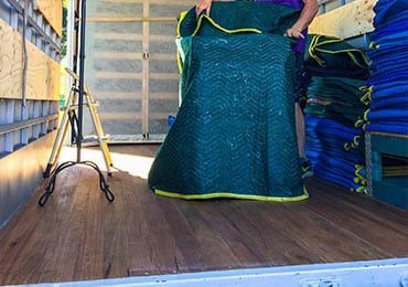 Man is Holding a Green Blanket Over a Piano in a Moving Truck — Bargara Removals in Rubyanna, QLD