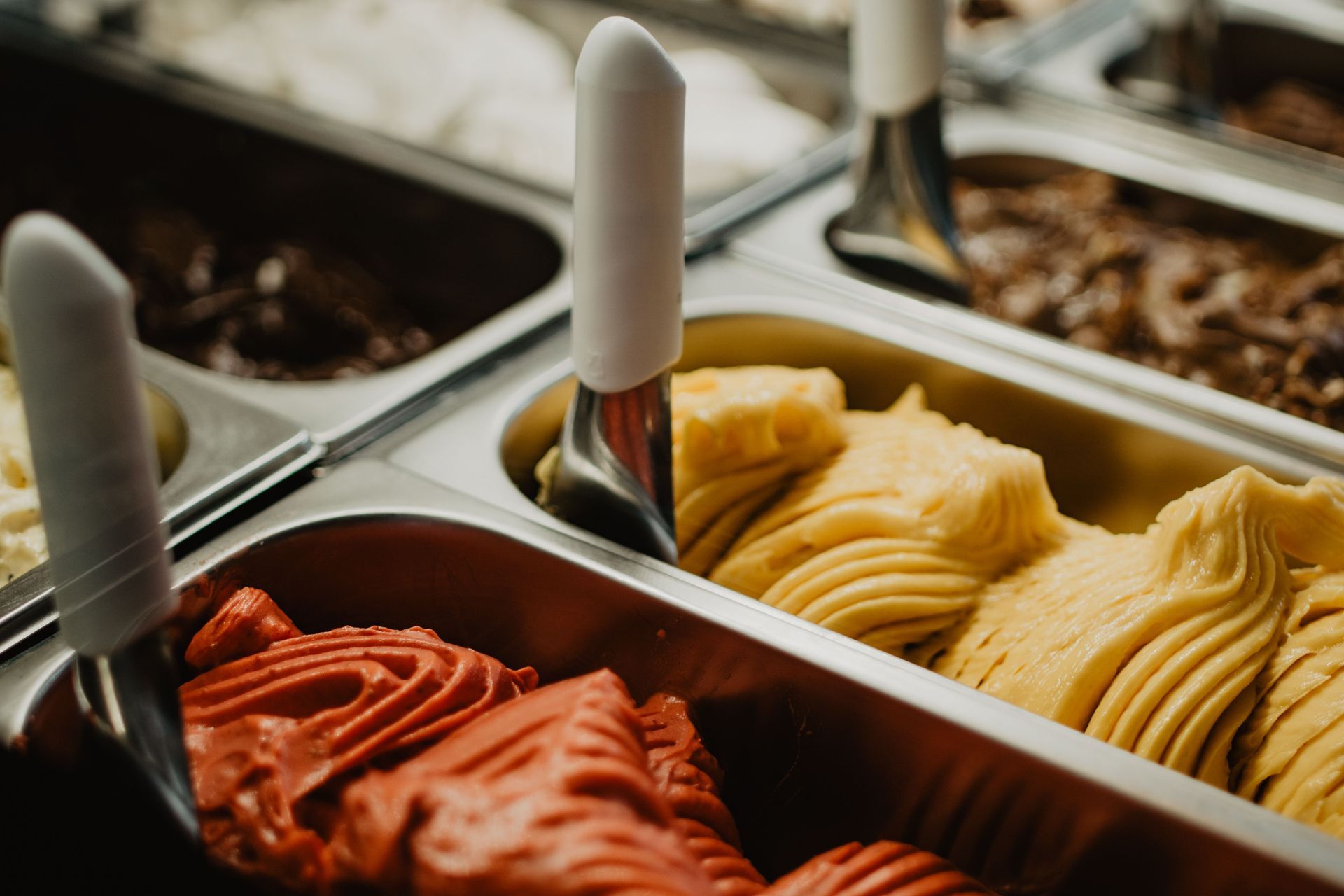 Bandejas de helado rojo, amarillo y blanco en una vitrina de postres con espátulas para servir.