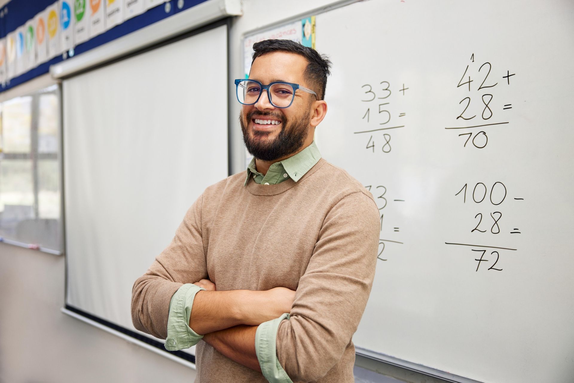 A teacher is standing in front of a whiteboard in a classroom with his arms crossed.