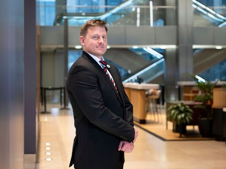 A professional man in a suit and tie stands with his hands clasped in a modern, well-lit office lobby.