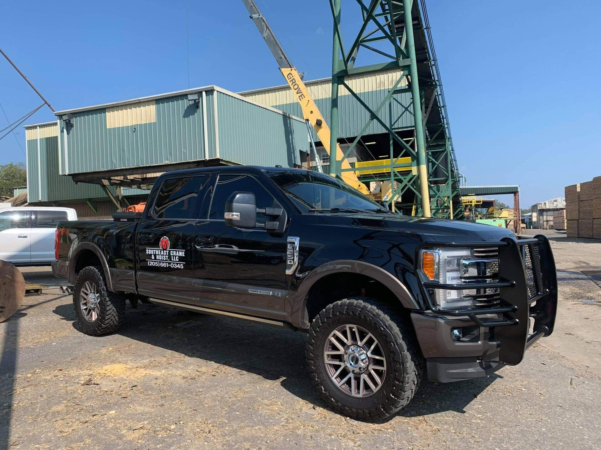 Black Ford F-350 pickup truck with a brush guard parked in front of a lumber mill with a crane.