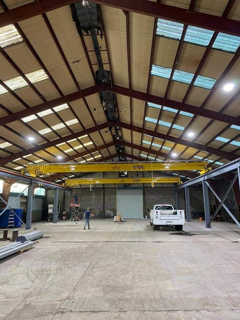 Inside a large industrial warehouse: a yellow overhead crane, a white truck, a person, and metal framing.
