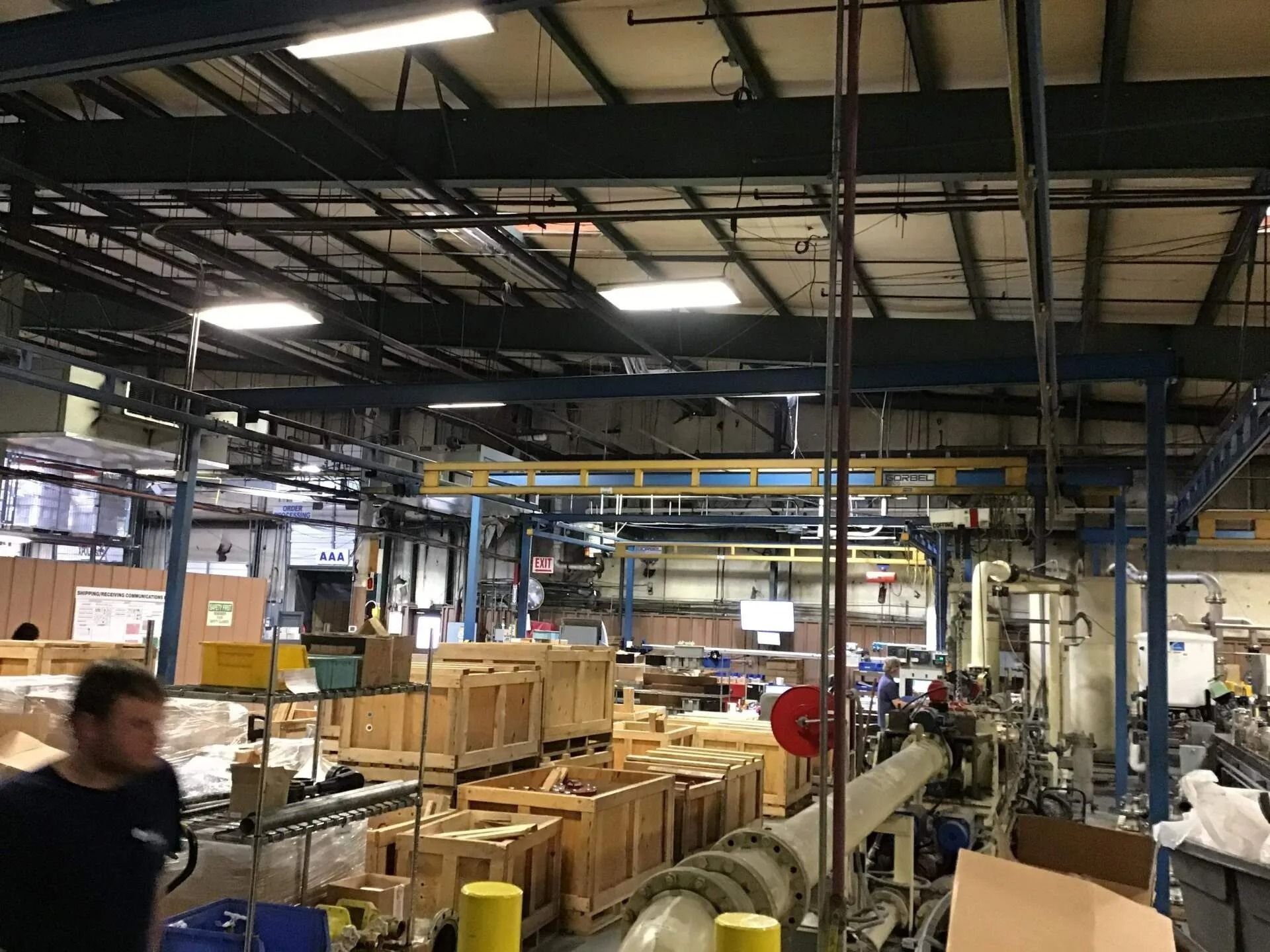 Industrial interior with crates, machinery, and a worker. Overhead beams and piping are visible.