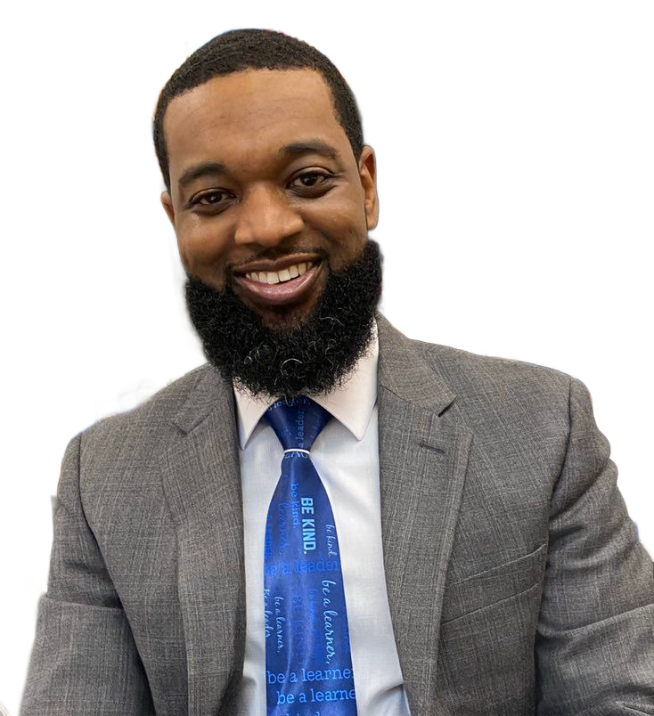 A man with a beard wearing a suit and tie is sitting in a classroom.