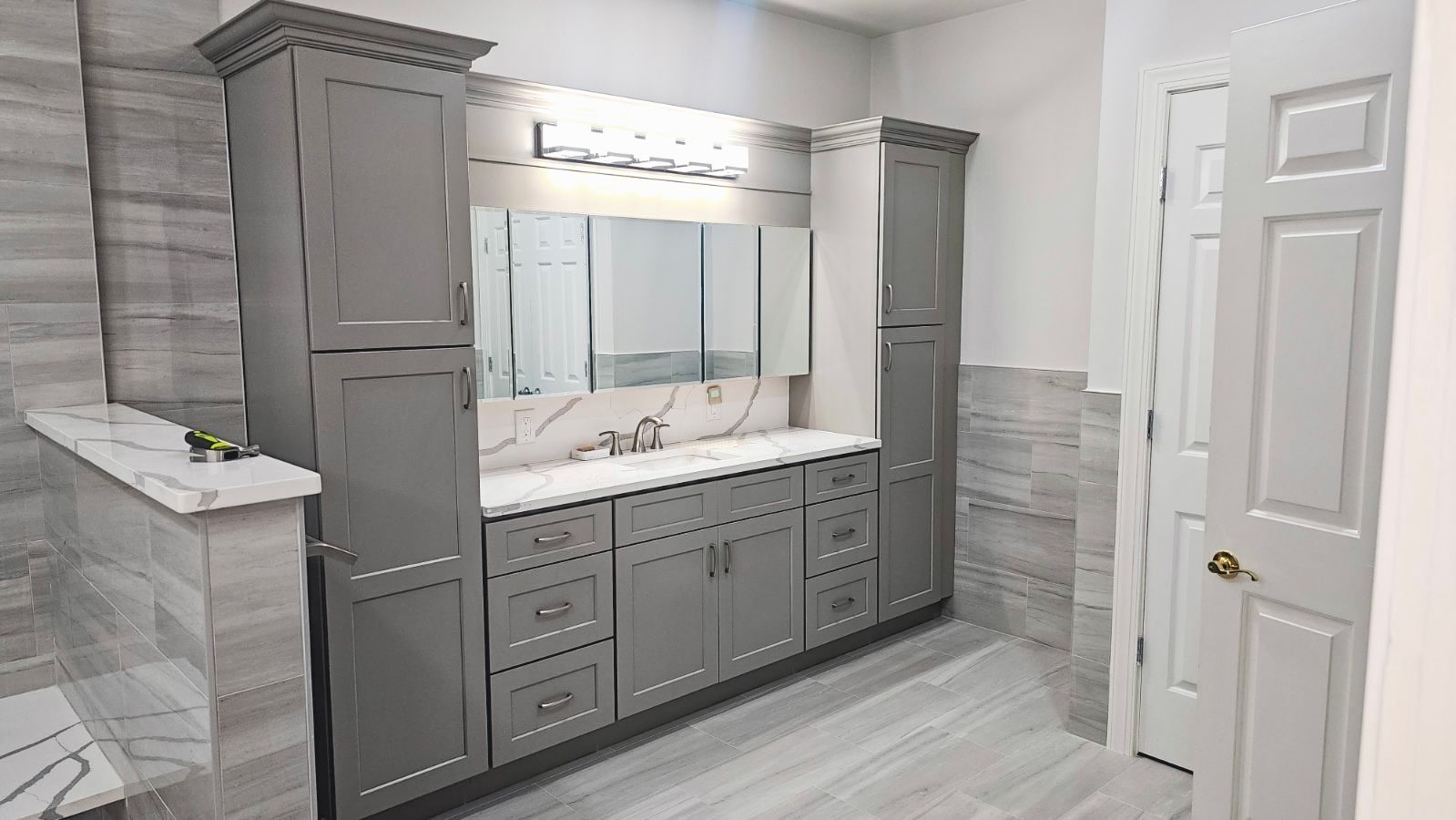 Gray bathroom with a vanity, large mirror, and tall cabinets. White countertop and light gray tile.