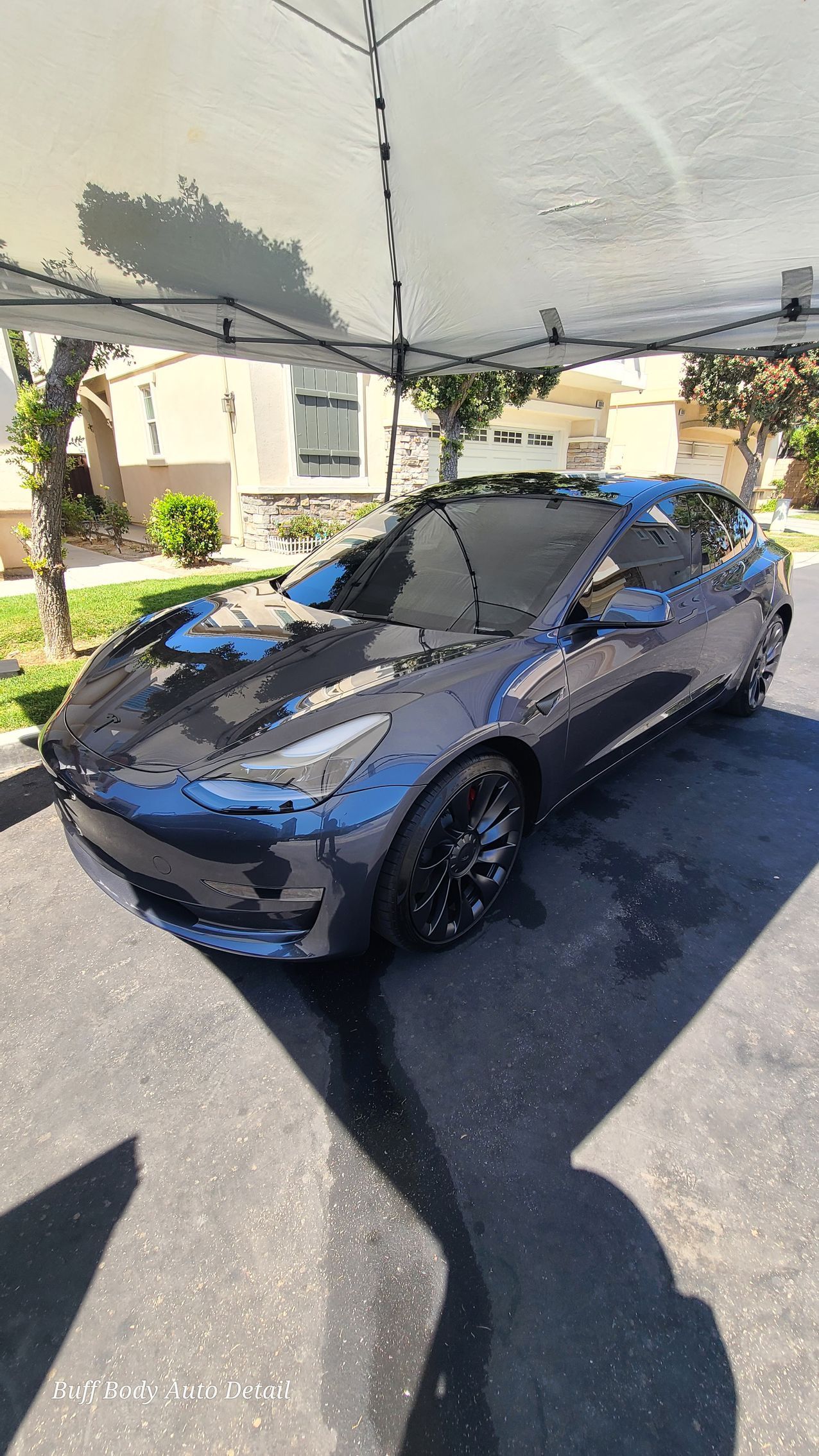 A tesla model 3 is parked under a tent in a driveway.