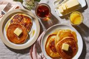 Pancakes with butter and syrup on plates, with a glass of juice on a breakfast table.