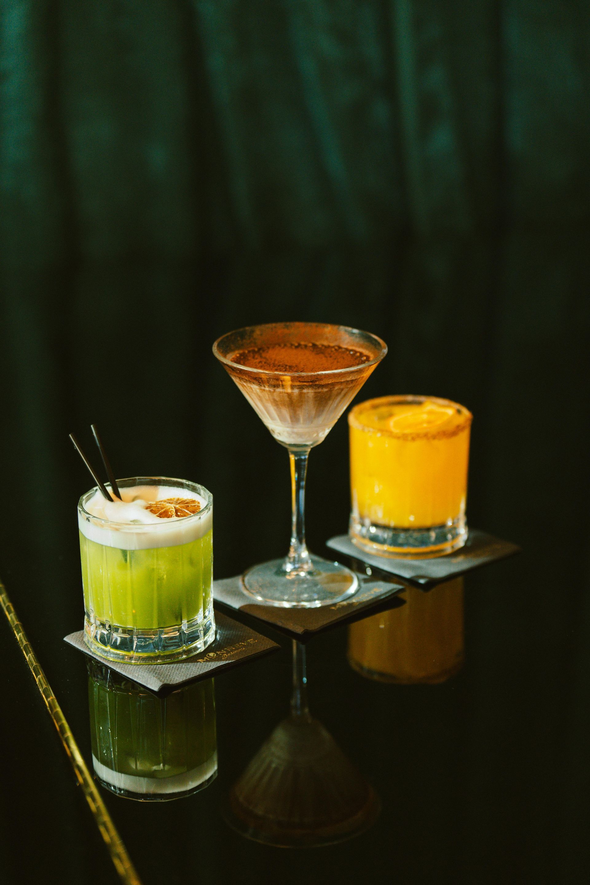 Three colorful cocktails on a glossy black table, with a dark green backdrop.