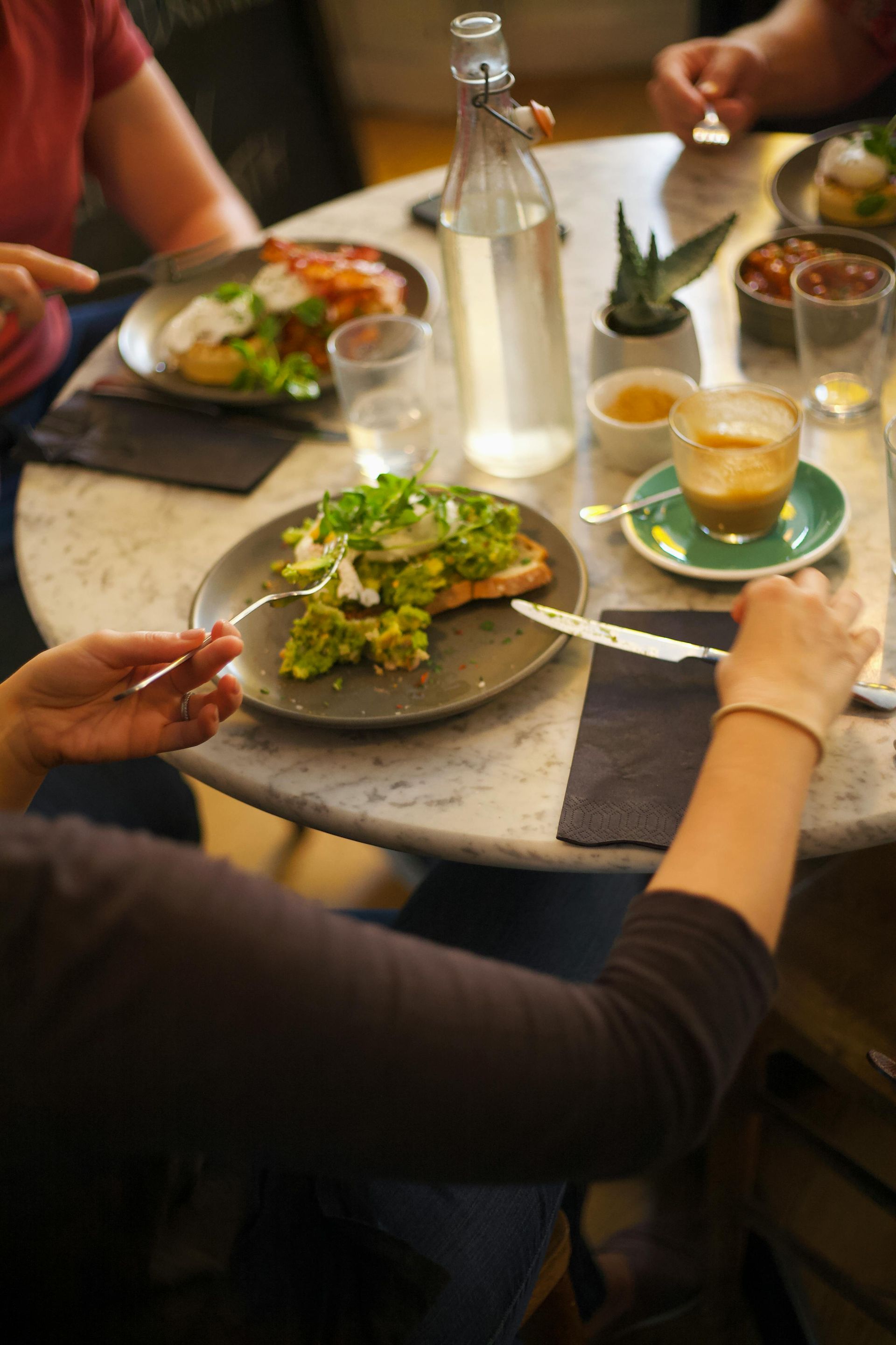 People sharing a meal at a round table with salads, drinks, and bread in a warm café setting