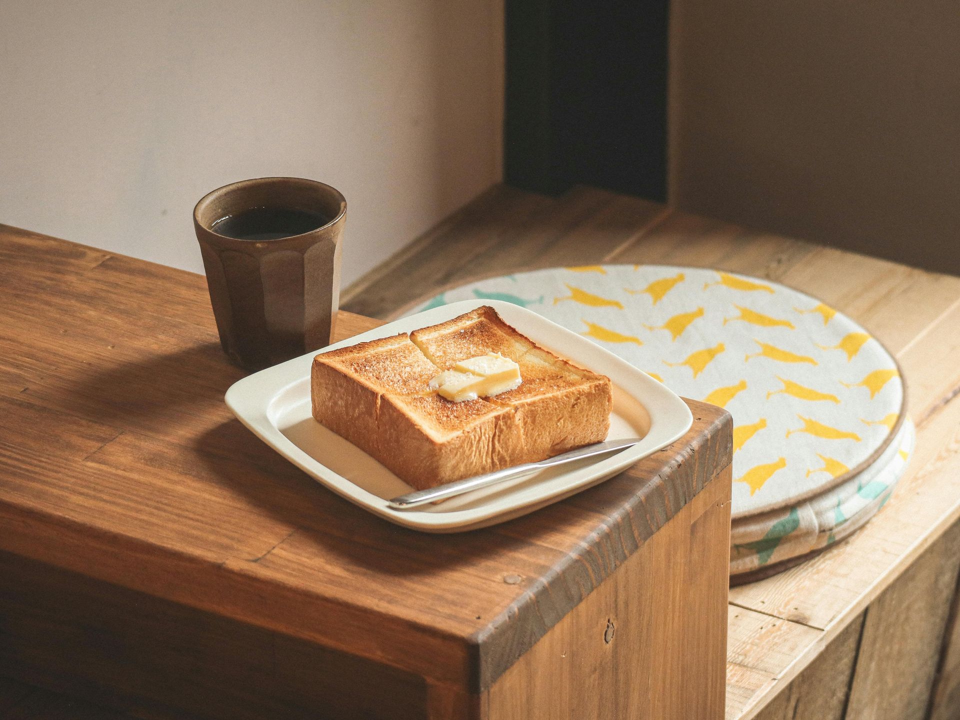 Toast on a plate beside a cup on a wooden table, with a patterned cushion in the background.