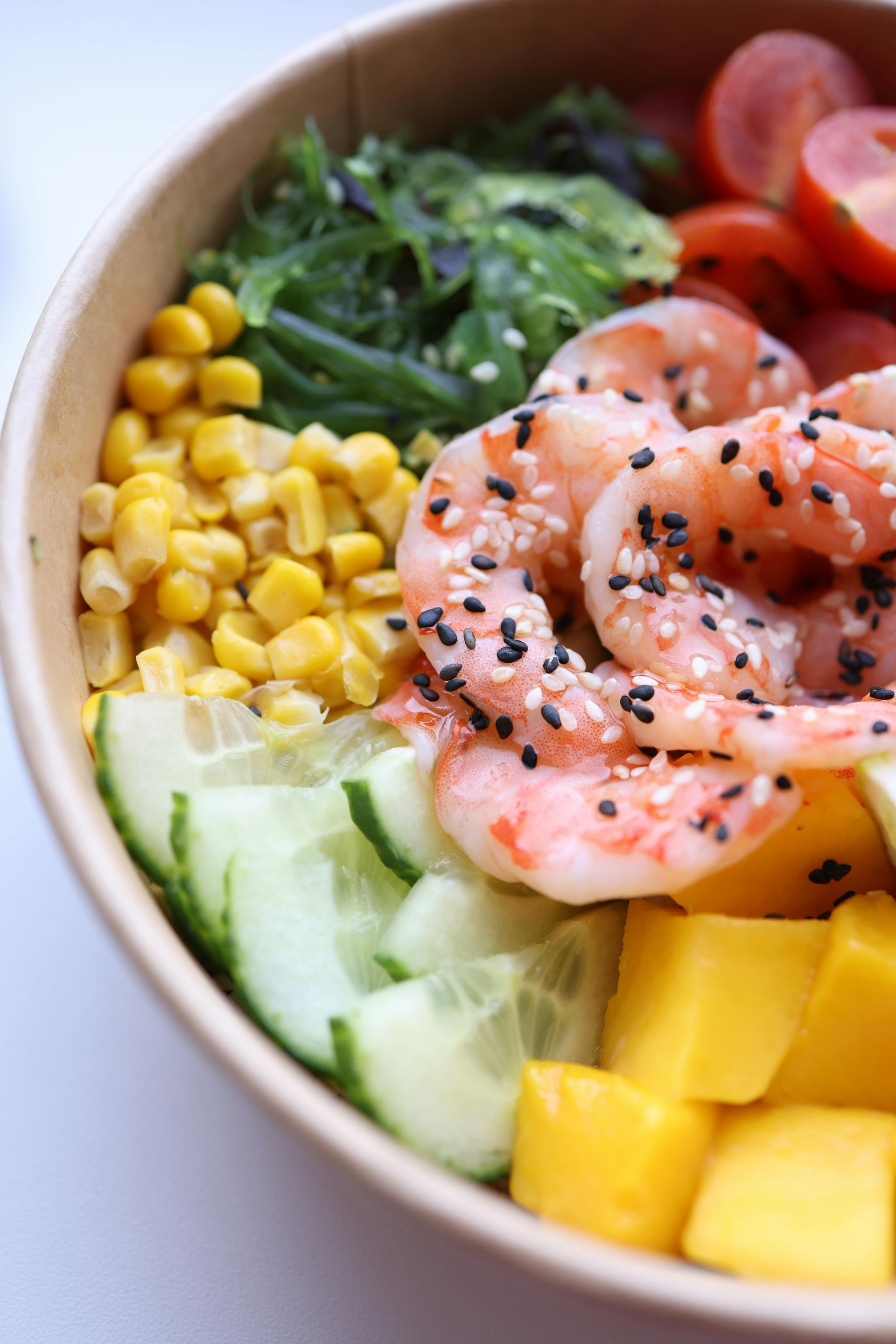 A colorful poke bowl with shrimp, corn, cucumber, mango, greens, and cherry tomatoes topped with sesame seeds