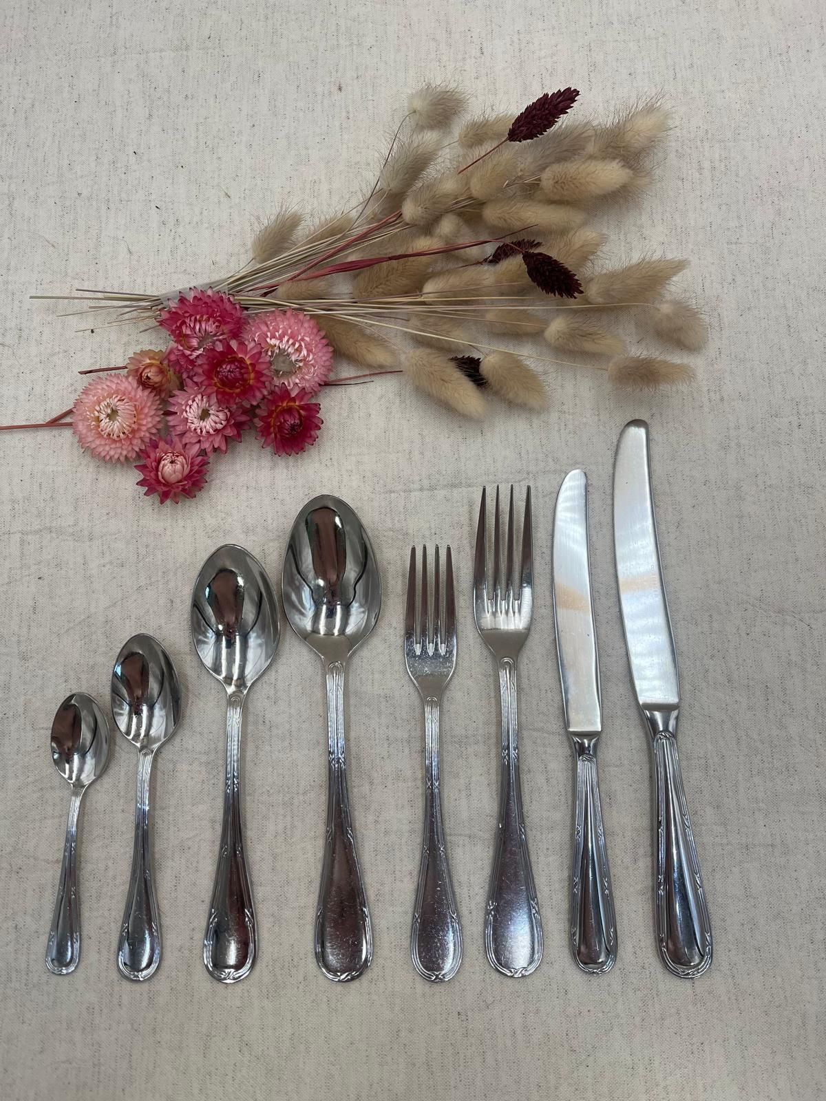 Cuillères, fourchettes et couteaux en argent disposés sous un petit bouquet de fleurs séchées sur un fond clair