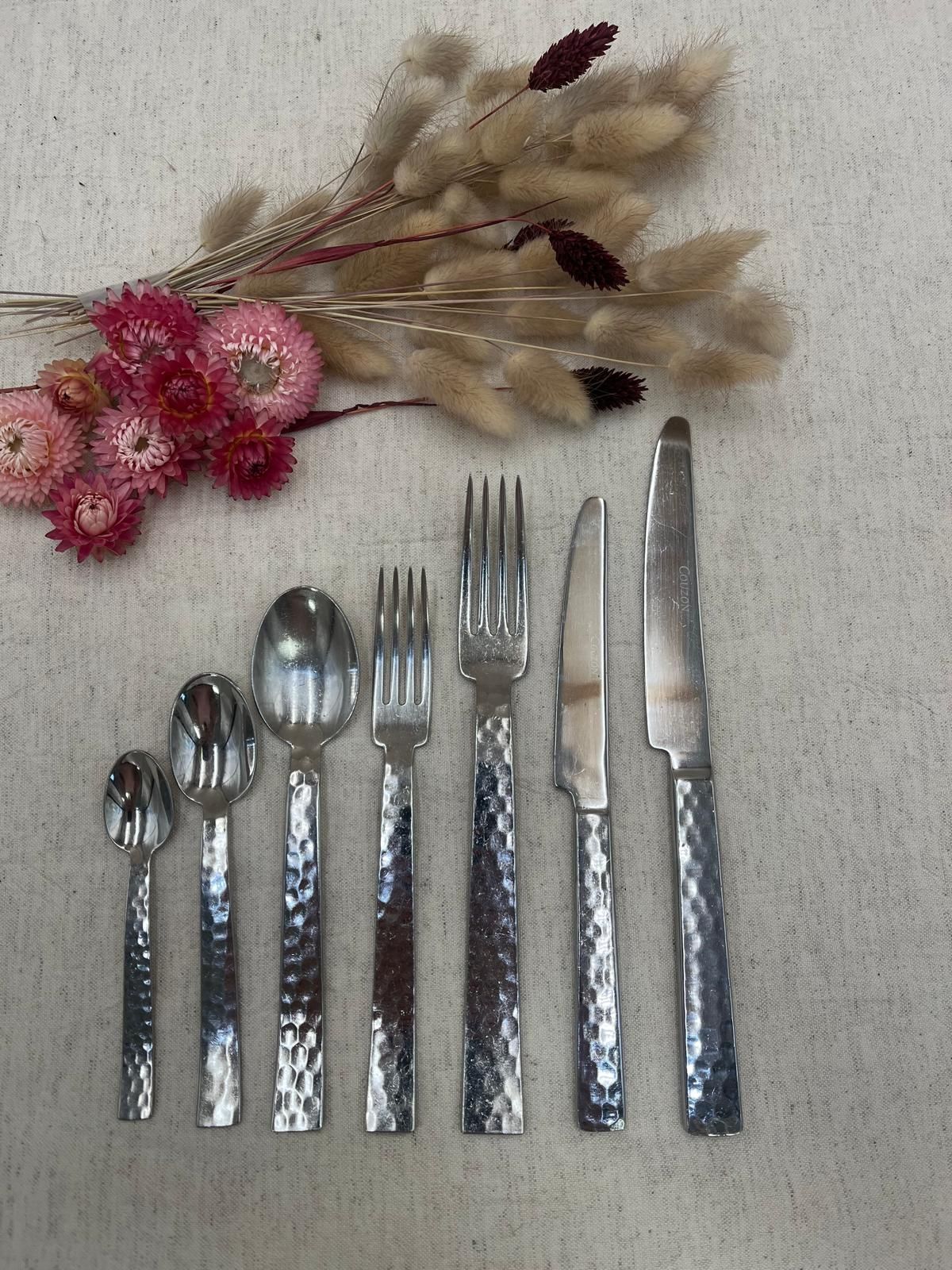 Sept ustensiles en argent disposés sous des fleurs séchées roses et beiges sur un fond clair