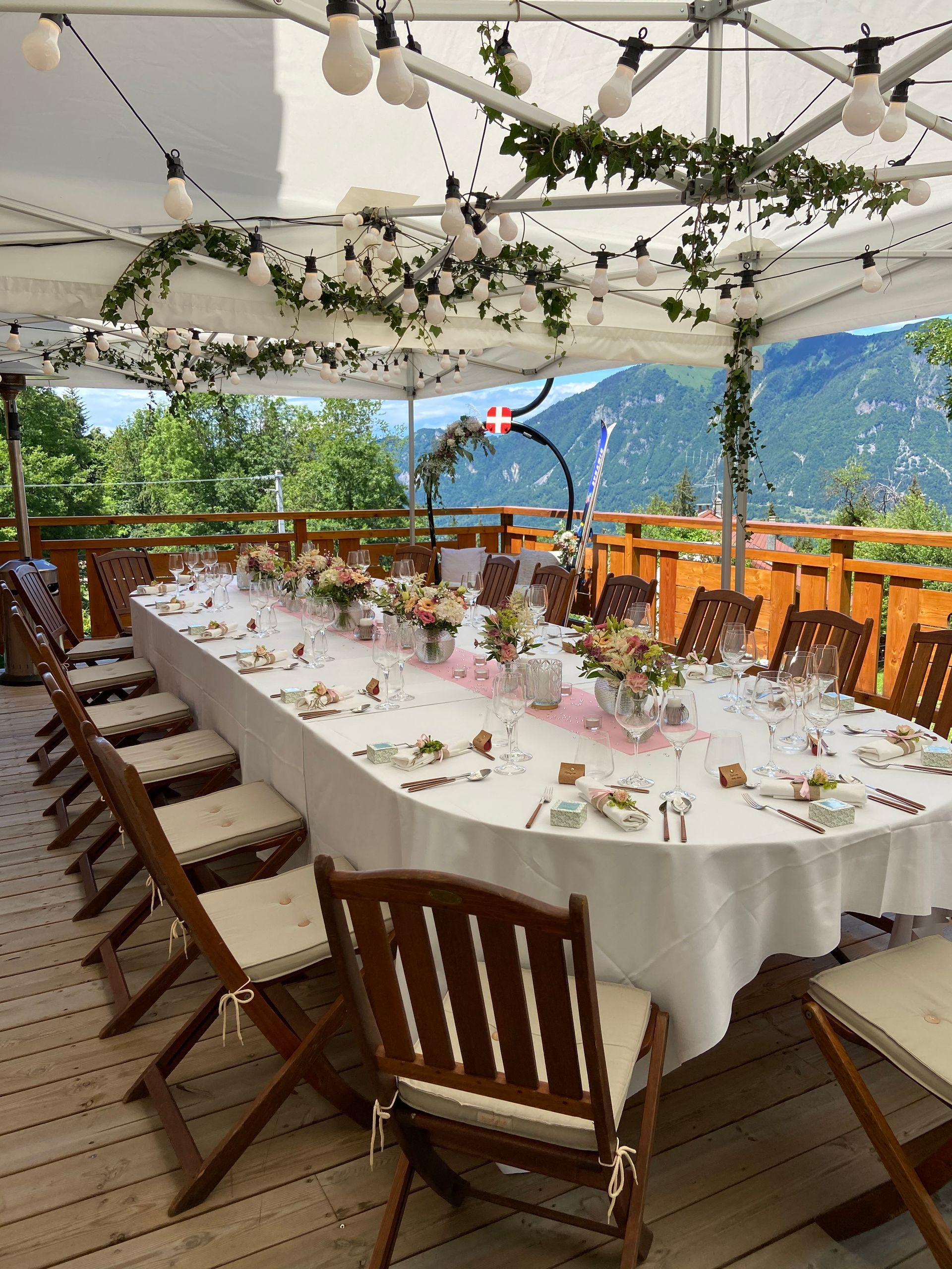 Longue table dressée pour un repas en plein air sous une canopée blanche, avec des fleurs et une vue sur les montagnes au loin.