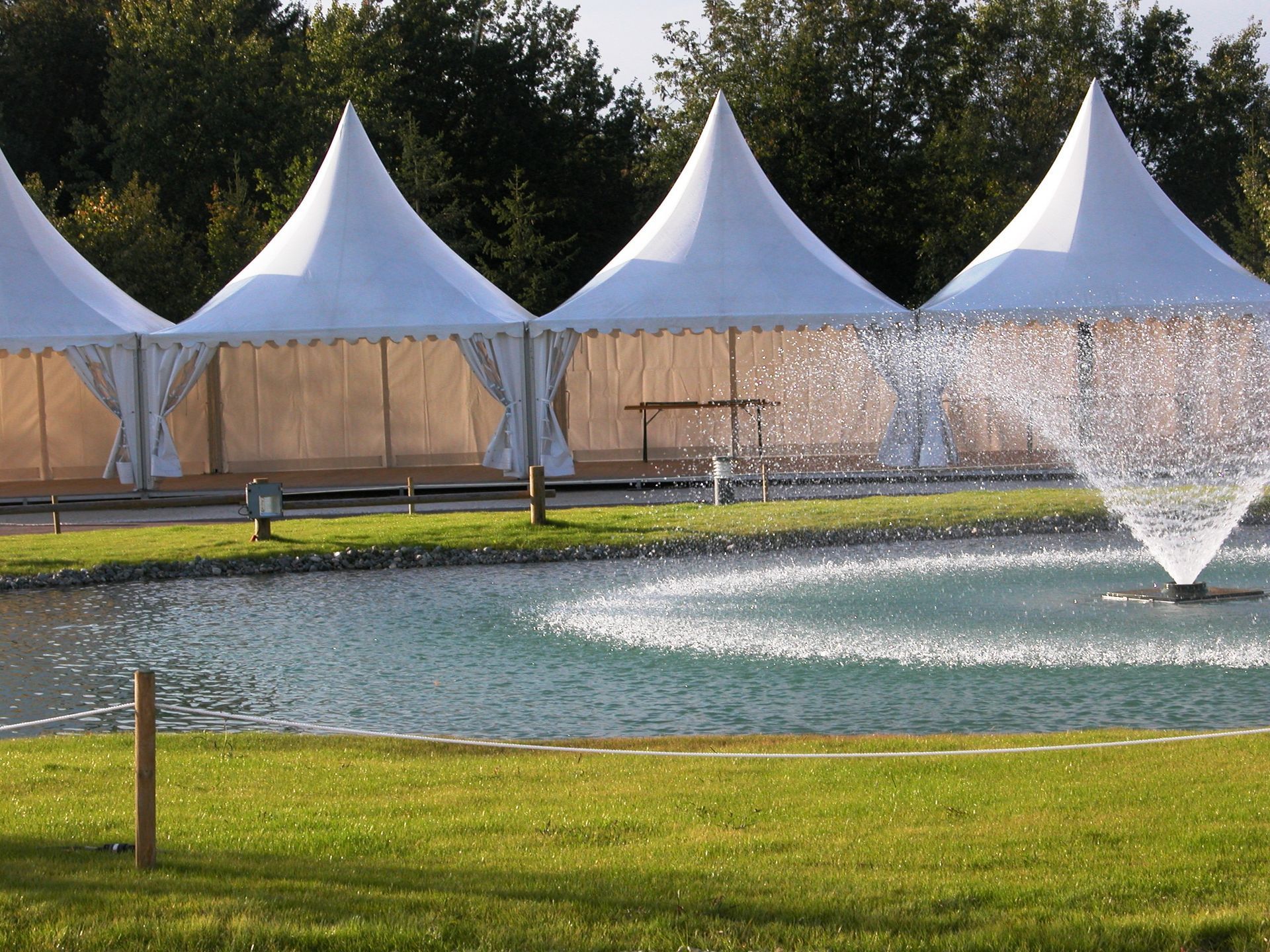Des tentes blanches près d'une fontaine jaillissant d'un étang dans un parc verdoyant