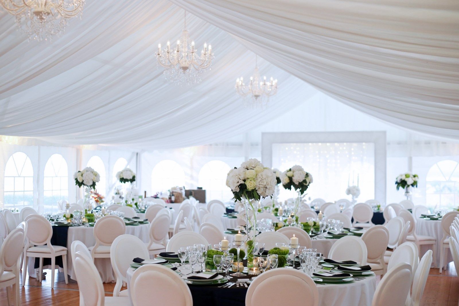 Élégante tente blanche pour réception de mariage, avec plafond drapé, lustres et tables décorées de fleurs