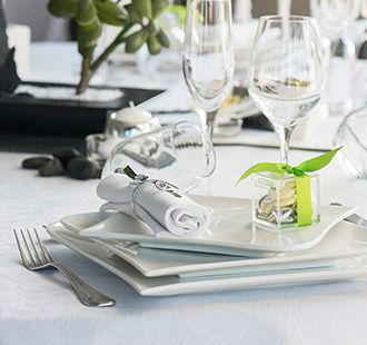 Élégante table blanche dressée avec assiettes empilées, serviette roulée, verres à vin et une petite touche florale verte.