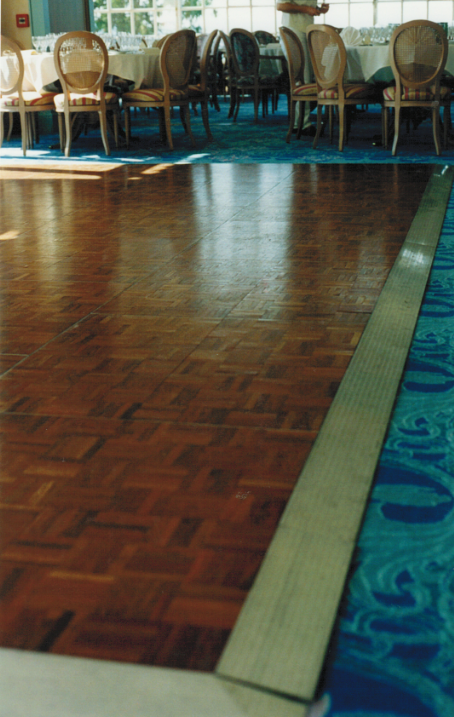 Piste de danse en bois avec tables et chaises en arrière-plan, bordée de moquette bleue.