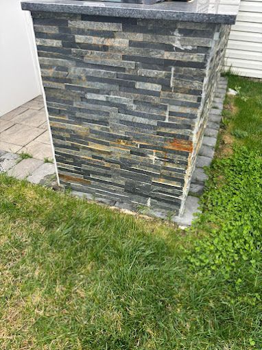 Stone-clad outdoor grill with dark gray and gold brick facade, set on a green lawn being repaired by melo powerwashing & painting in linwood, nj