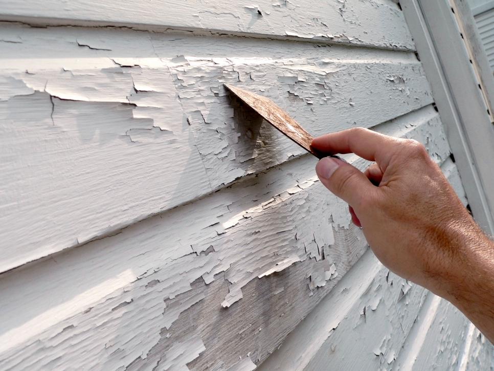 A person is peeling paint off of a white siding with a spatula