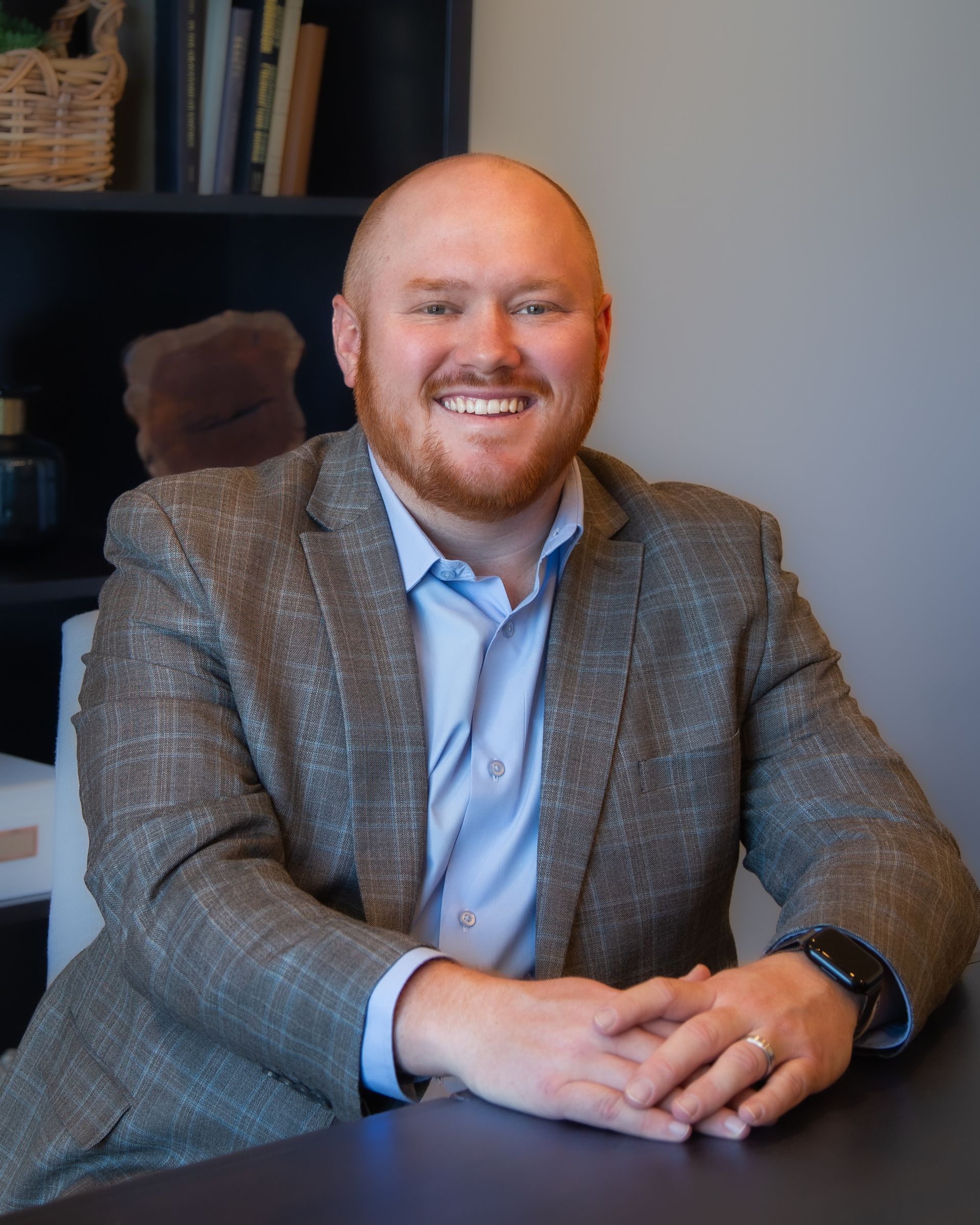 A man in a suit is sitting at a table with his hands folded and smiling.