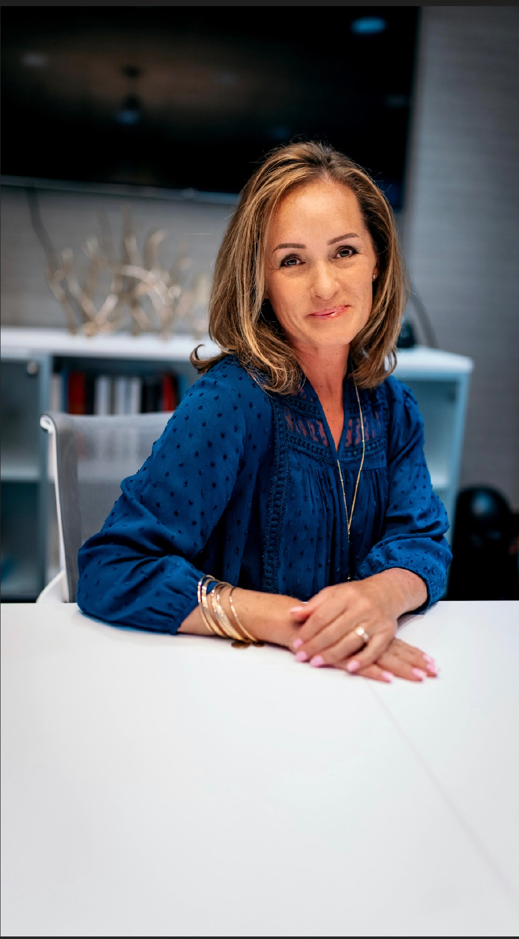 A woman in a blue shirt is sitting at a table with her hands folded.